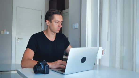 Young Photographer Using Laptop Processing Images on His Laptop and Drinking Coffee at Home