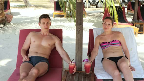 Couple Lies on Sunbeds and Drinks Tasty Beverages Under Shed