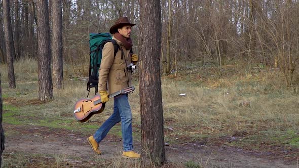 A Backpacker with a Guitar and a Camera Walks Along a Trail in the Forest