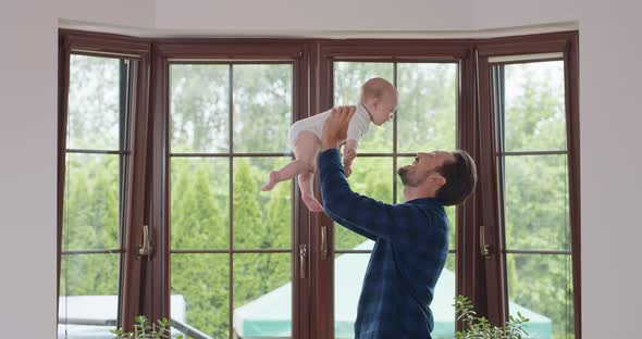 Bearded and Mustashed Father Holds on Raised Hands His Adorable Baby and Talks to It Window and a