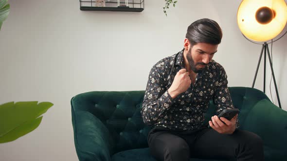 Latin Handsome Bearded Man Sitting on a Greencolored Couch Looking at His Mobile Phone and Cheering