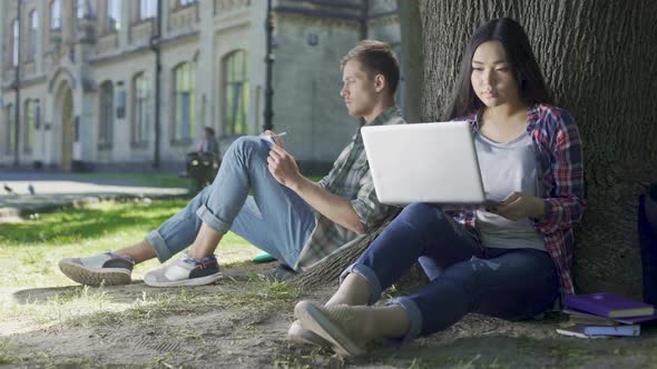 Young Woman Using Laptop and Guy Taking Notes in Notebook, Sitting Under Tree