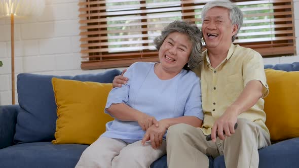 Smiling Asian senior couple sitting on a sofa in the living room and watching TV at home.