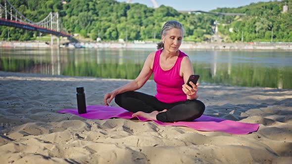 Elderly Woman Takes Selfie Sitting on Pink Mat on River Bank