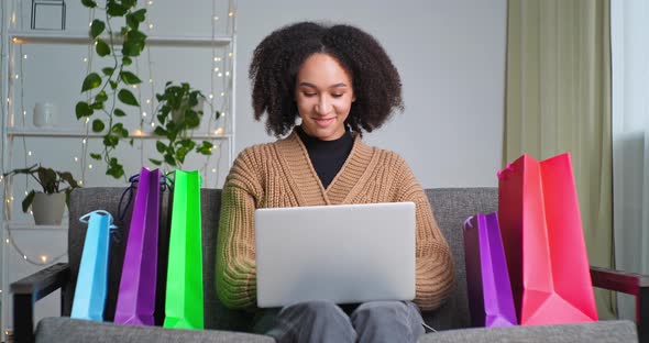 Successful Ethnic Gambling Girl Sitting on Sofa Surrounded By Shopping Bags Typing on Laptop Wins