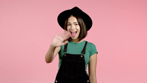 Young Mixed Race Friendly Woman in Hat, Glasses and Overall Waving Hand - Hello. Greeting, Say Hi To