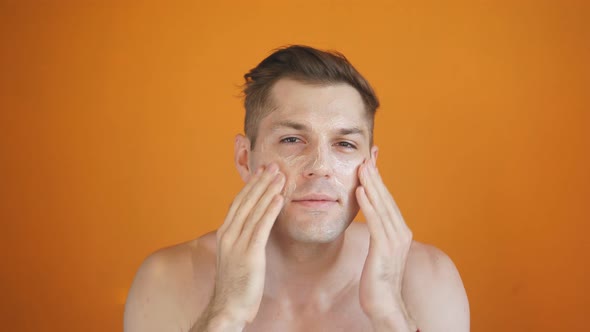 Caucasian Man Takes Care of His Face Skin, Moisturizes It with Cosmetic Cream, Isolated Orange