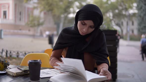 An Attractive Muslim Girl Writes in a Notebook Sitting in a Cafe with a Book