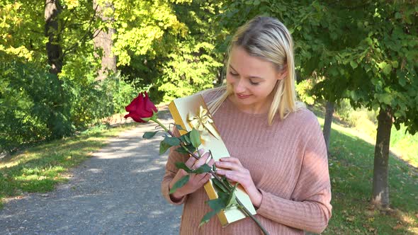 A Young Beautiful Woman Holds a Rose and a Present and Smiles Happily at the Camera in a Park