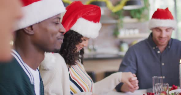 Happy group of diverse friends in santa hats praying, celebrating meal at christmas time