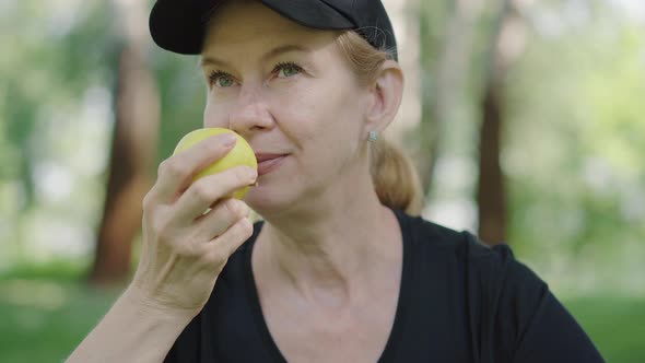 Close-up of Happy Caucasian Woman in Sportswear and Cap Smelling Tasty Apple in Summer Park