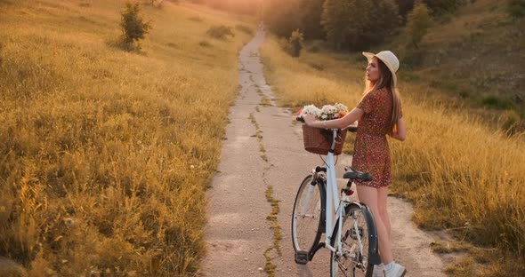 Smiling Happy Girl in Dress and Hat Riding Retro Bicycle in the Park and Looking at Camera