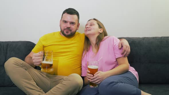 Young Couple of People are Sitting on Sofa and Drinking Beer From Transparent Glass Glasses