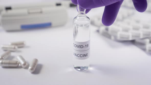 A hand in protective gloves picks up an ampoule of medicine from a white table 