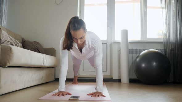 Woman Stands in Plank Pose Watching Tutorial on Mat in Room