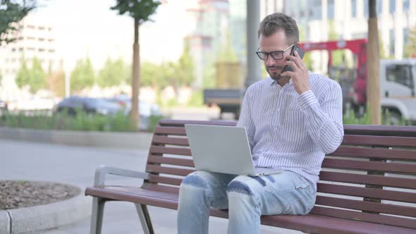 Angry Young Adult Man Talking on Phone and Using Laptop While Sitting Outdoor on Bench