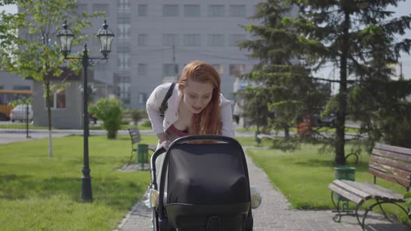 Young Happy Redhead Mother Walking with Baby Carriage and Smiling Along the Street on a Nice Spring