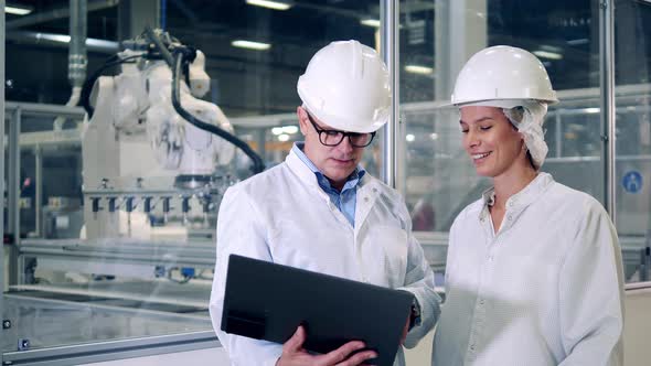 Two Engineers in Hardhats are Talking Next to Robotic Machines