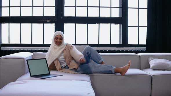 Young Muslim Girl Sits on Couch and Looks at the Laptop in Bright Beige Studio