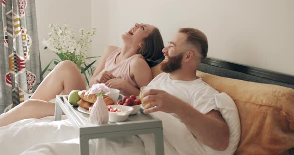 Cheerful Couple Having Breakfast in Bed and Laughing While Looking at Each Other in Morning, Young