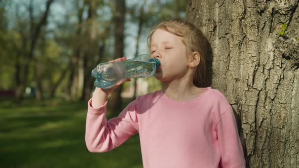 Thirsty Blonde Girl Drinks Water Near Old Tree in City Park