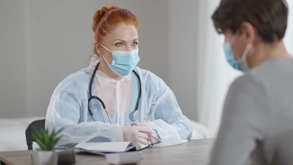 Portrait of Professional Female Doctor in Covid Face Mask Consulting Male Patient on Coronavirus