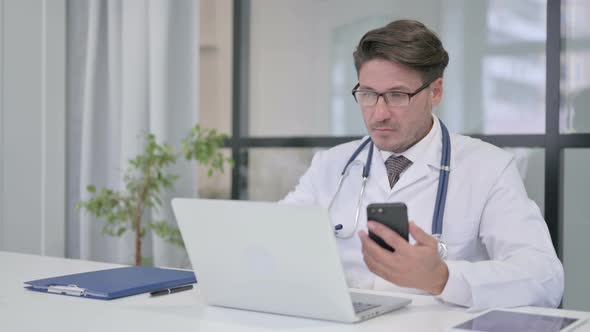 Doctor using Smartphone while Working on Laptop in Clinic