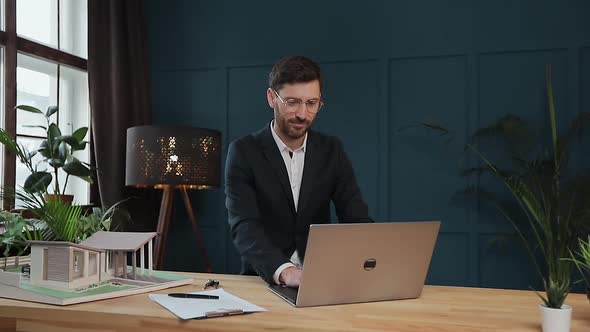 Male Designer in Office Working on Laptop with Model House.