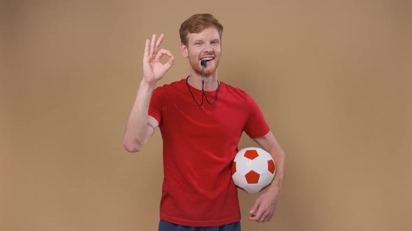 Unny Man on a Beige Studio Background Coach with a Ball and a Whistle Shows an Approval Sign Ok