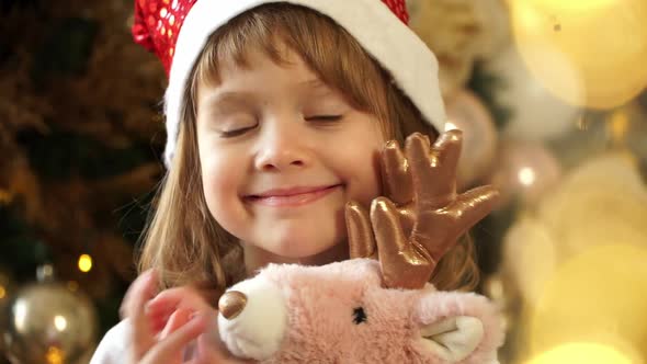 Little Girl in Red Santa Hat Looks at Camera Dances and Smiles on Background Christmas Tree