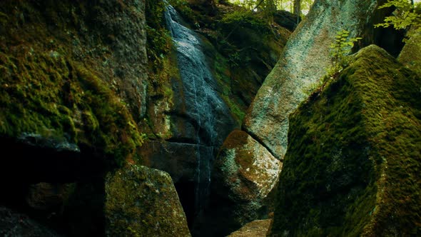A small waterfall cascades down a ledge surrounded by large moss covered rocks.