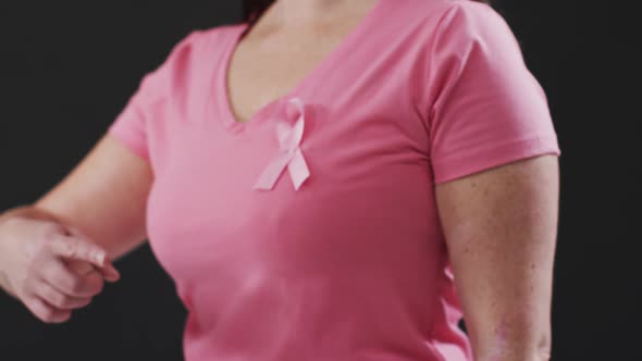 Mid section of a woman pointing to the pink ribbon on her chest against black background