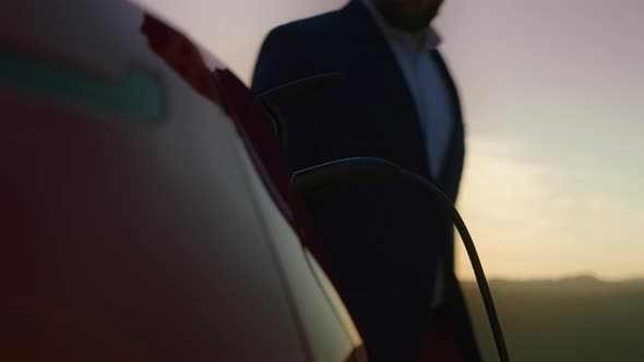 Handheld Shot of Caucasian Man in Black Suit Charging Electric Car at Sunset