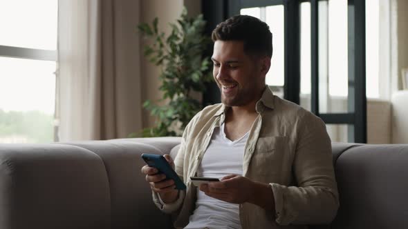 Positive man sitting on sofa in apartment making online payment. 