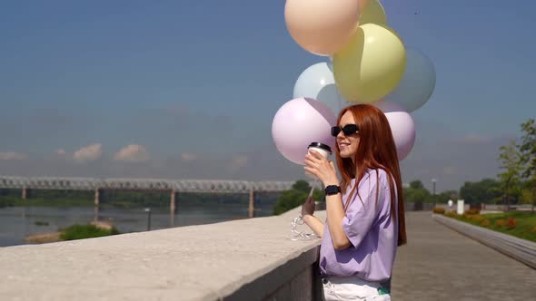 Happy Redhaired Girl Wearing Sunglasses Holds Helium Balloons and Drinks Coffee on Riverside