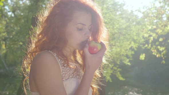 Portrait of Beautiful Slim Caucasian Woman with Red Hair Eating Apple Standing in the Sun Light in