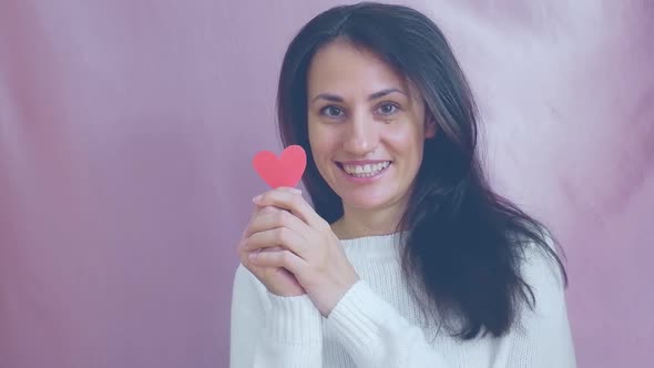 Smiling Funny Young Brunette in a White Sweater Smiles Blinks and Waving Red Hearts on a Pink