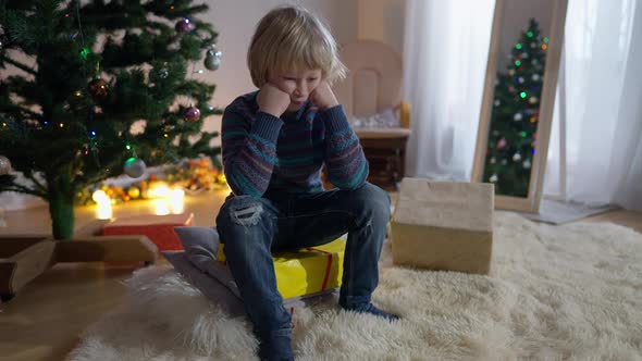 Wide Shot Sad Cute Little Boy Sitting on Christmas Gift Box at New Year Tree in Living Room Thinking