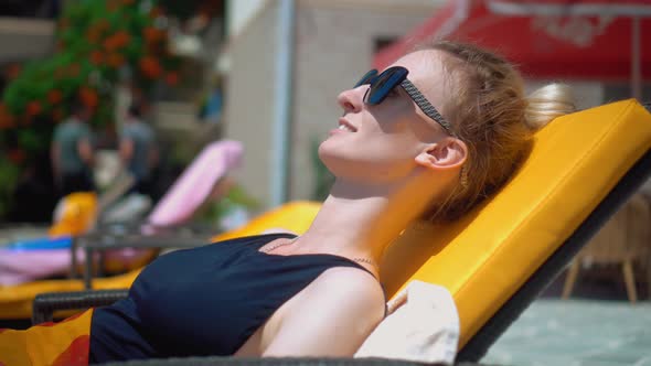 Woman in sun protective glasses and black swimsuit lies on sun lounger by pool,