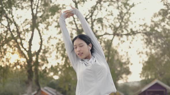 Asian woman athlete listening music wearing sportswear stretching with arms raised.