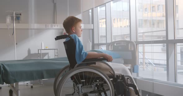 Side View of Pensive Kid Sitting on Wheelchair Looking Out of Window in Hospital Ward