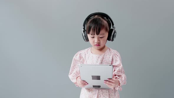 ittle Asian girl with headphone enjoy watching video online on tablet and dancing happily.