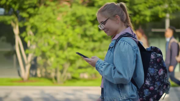 Cheerful Caucasian Female Student Using Cellphone Walking with Backpack Outdoors