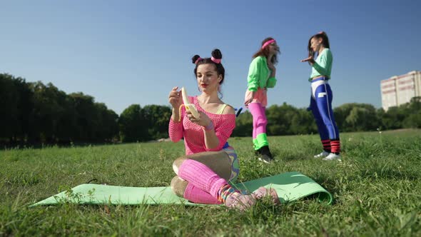 Wide Shot Happy Woman Peeling Banana Sitting on Exercise Mat with Blurred Women Laughing Pointing