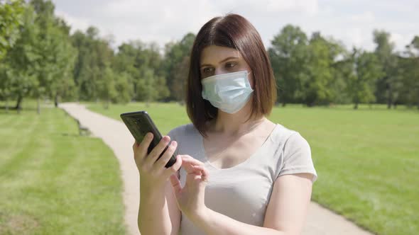 A Young Caucasian Woman in a Face Mask Works on a Smartphone on a Pathway in a Park on a Sunny Day