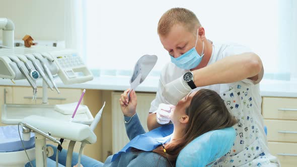 A male dentist in rubber gloves with a medical tool treats a girl's teeth in a clinic.