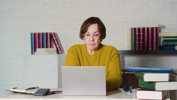 Dolly Shot of an Elderly Caucasian Woman in Glasses Working in Front of a Laptop Monitor