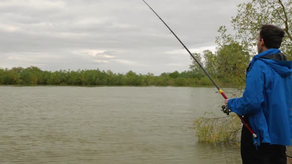 An angler in a blue jacket with a long fishing rod on the bank of a river