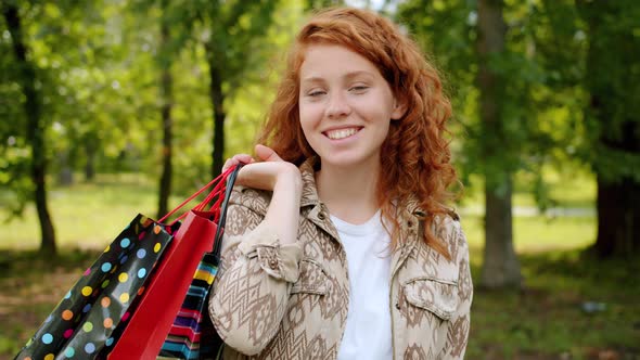 Portrait of Cheerful Teenager with Paper Bags Standing Outdoors in Park Smiling