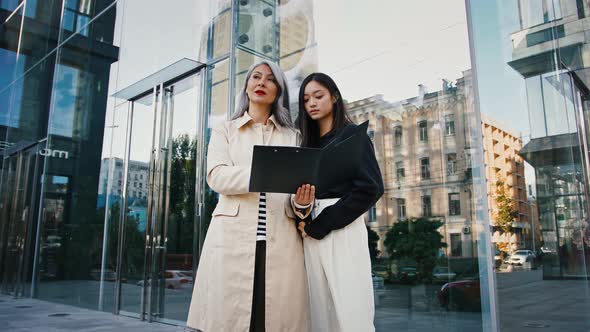 Asian Grayhaired Lady Holding Folder and Discussing Details of New Project with Her Young Colleague